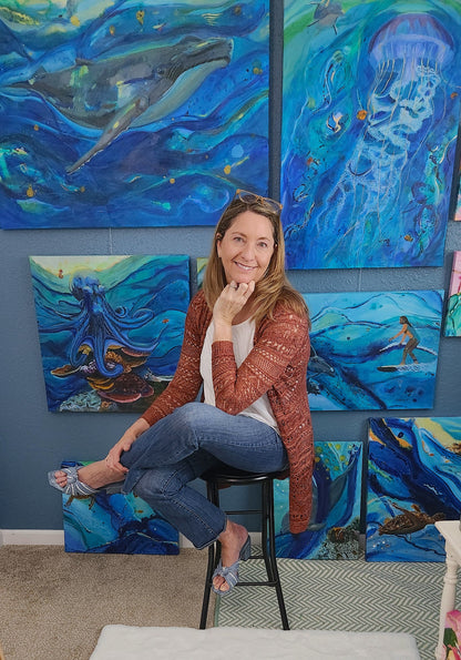 A woman sits on a black stool in front of a blue wall decorated with multiple colorful paintings of ocean life, including whales, jellyfish, octopus, and a surfer. She is wearing a rust-brown cardigan over a white top, blue jeans, and blue sandals.