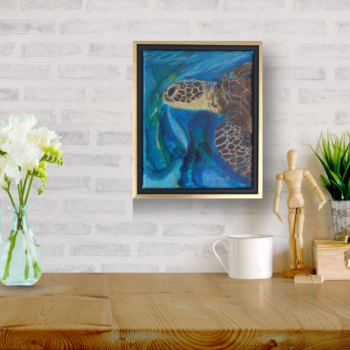 A framed abstract painting of a sea turtle in shades of blue, green, and brown, hanging on a white brick wall above a wooden table with a vase of white flowers, a white mug, and a wooden artist mannequin.
