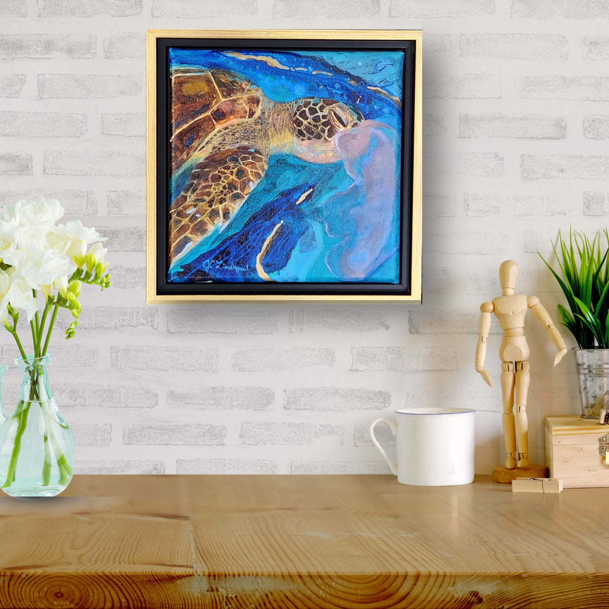 A framed abstract painting of a sea turtle hangs on a white brick wall above a wooden table with a vase of white flowers, a white mug, a small wooden figurine, and a potted plant.
