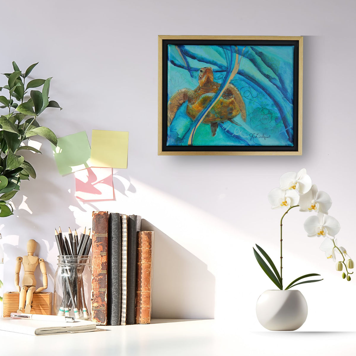 A framed painting of a sea turtle in blue water hangs on a white wall above a styled desk with books, a plant, sticky notes, and a white orchid vase.