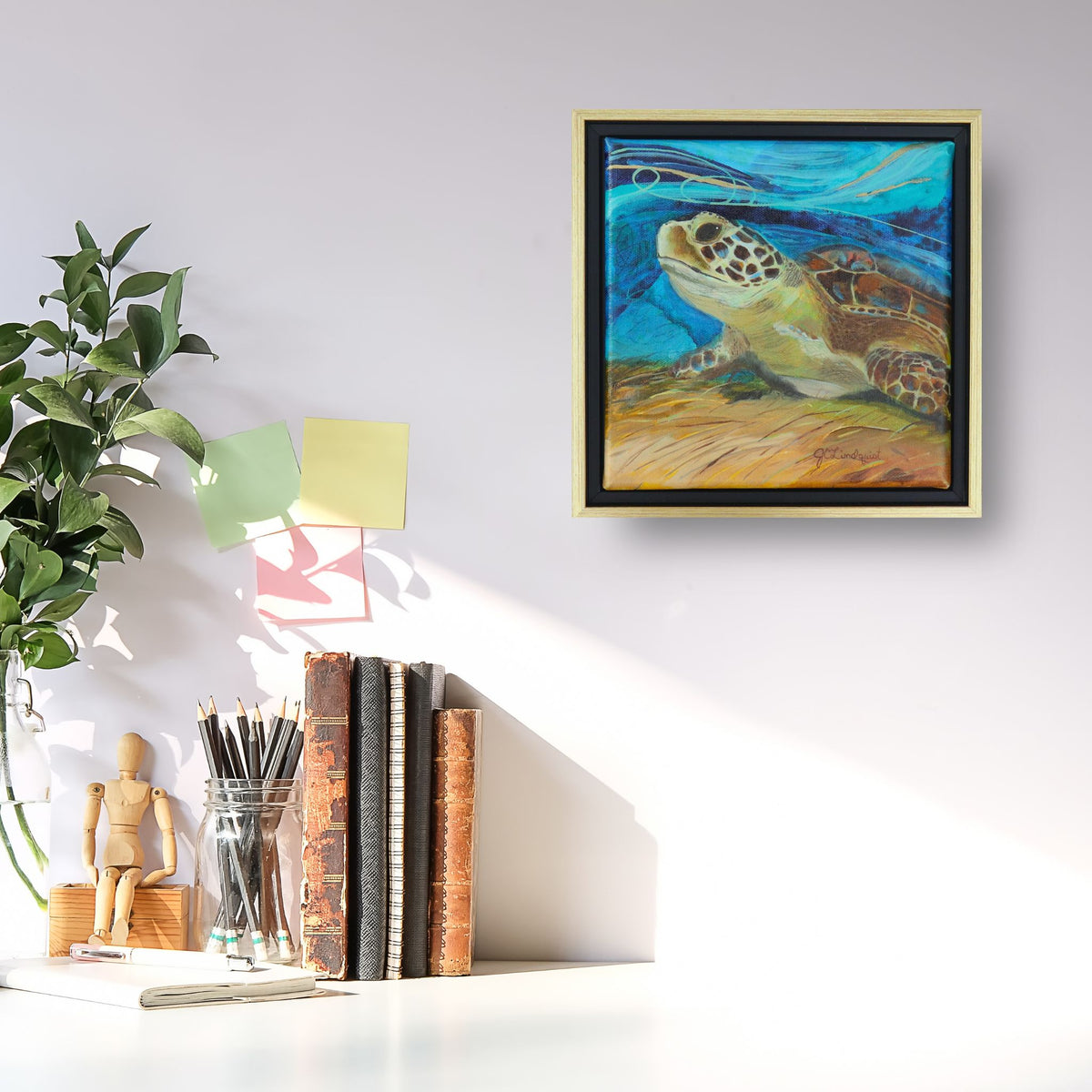 Picture of a white desk with books, pencils and branches as decor.  A square painting of a sea turtle with a blue abstract background is displayed inside a black and gold floating frame.