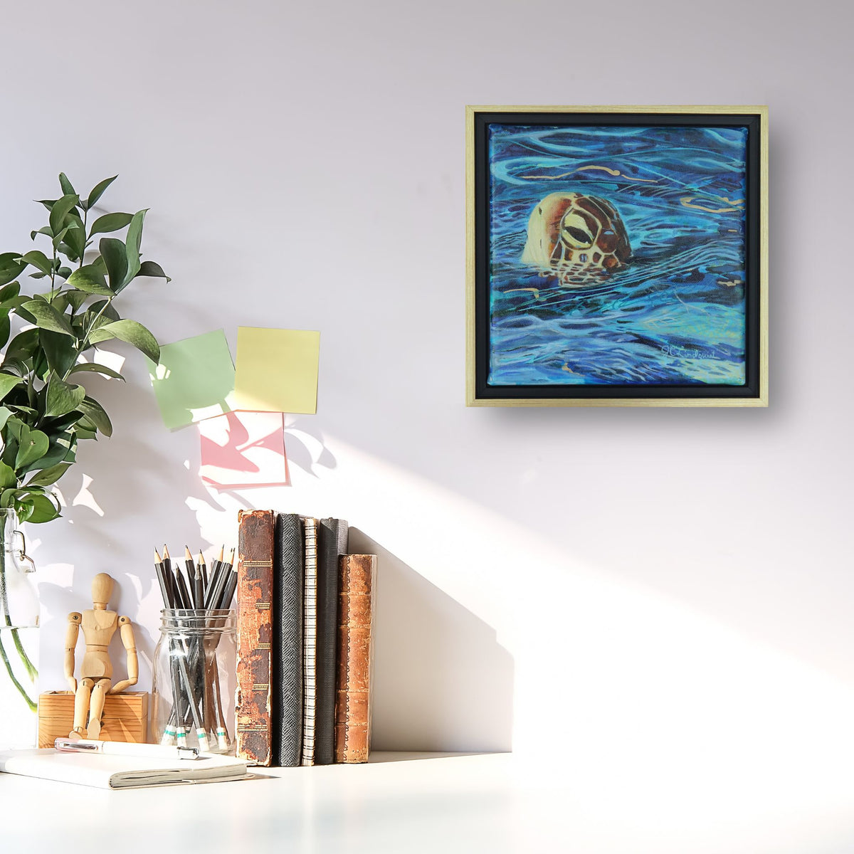 A framed square painting of a sea turtle swimming in blue water, displayed on a white wall above a desk with books, pencils, and a plant.