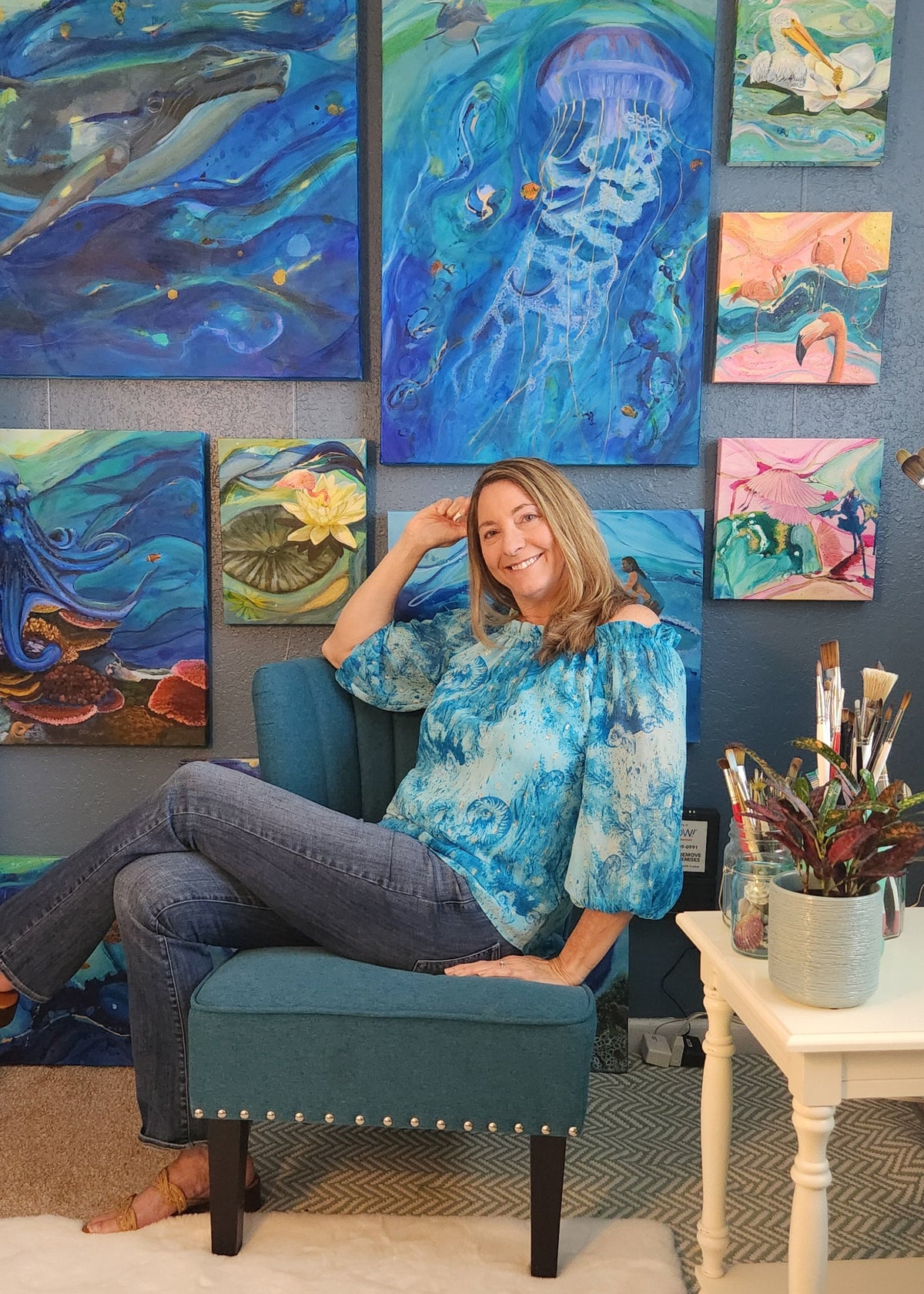 A woman sits cross-legged on a teal upholstered bench in front of a blue wall decorated with multiple colorful abstract paintings. She is wearing a blue patterned off-the-shoulder blouse and jeans.