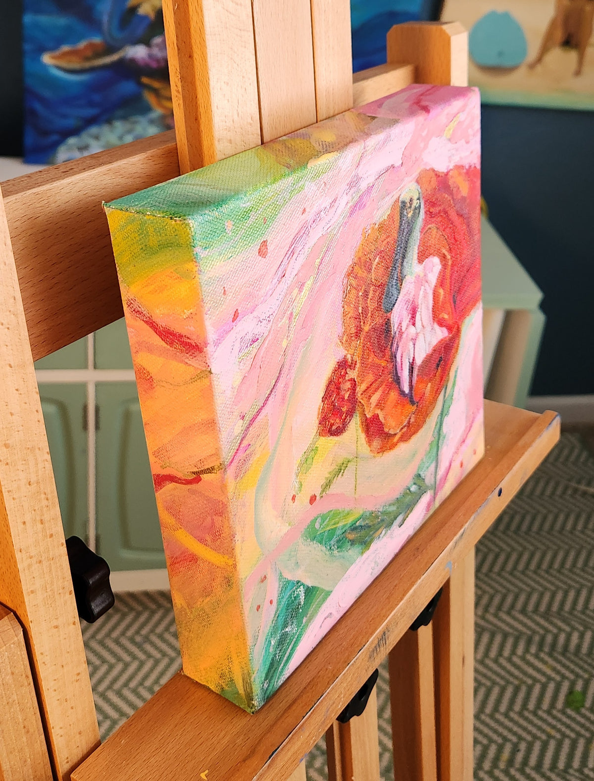 A colorful abstract canvas painting with pink, orange, yellow, green, and white brushstrokes, displayed on a wooden easel in an indoor setting.
